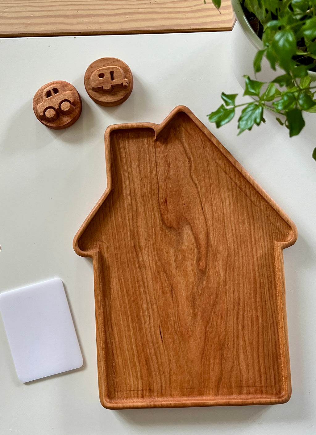 Wooden tray shaped like a house with two small round wooden toy pieces and a white rectangular card nearby on white surface
