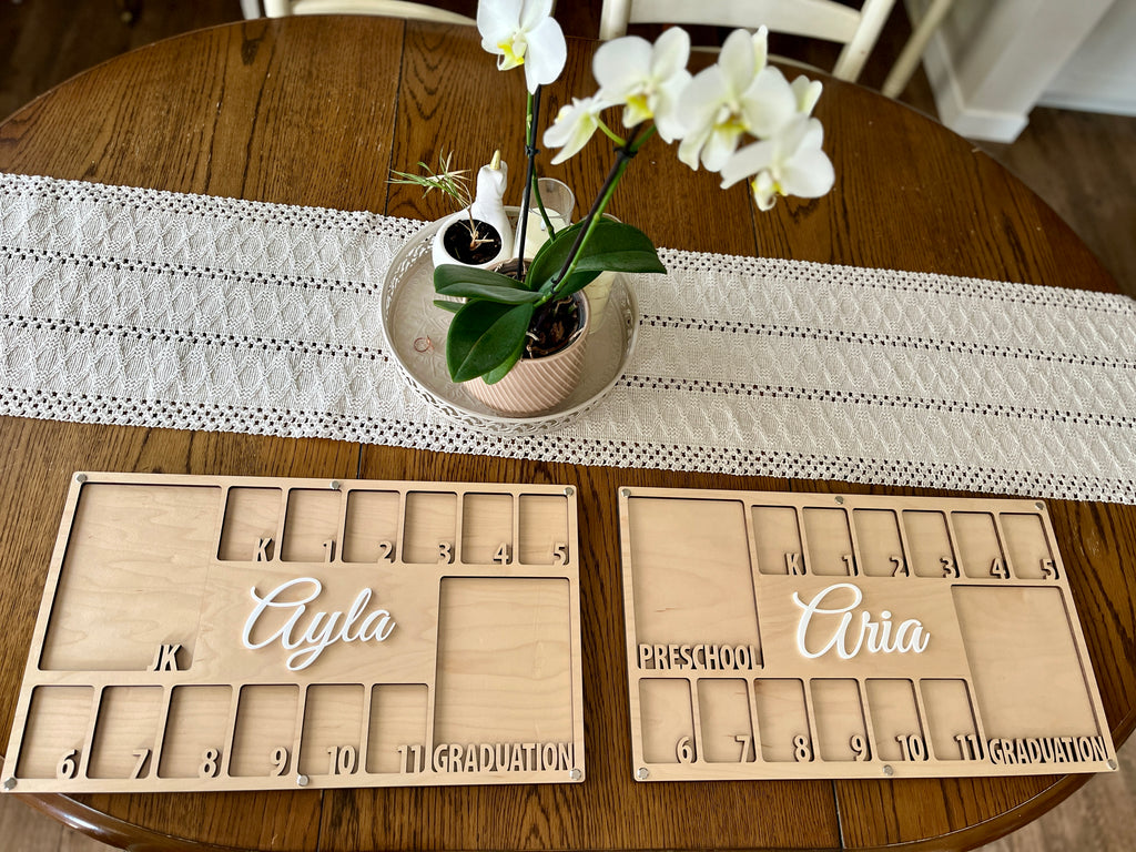 Two wooden milestone boards with engraved numbers and white script names displayed on a wooden table with a lace runner and white orchids centerpiece