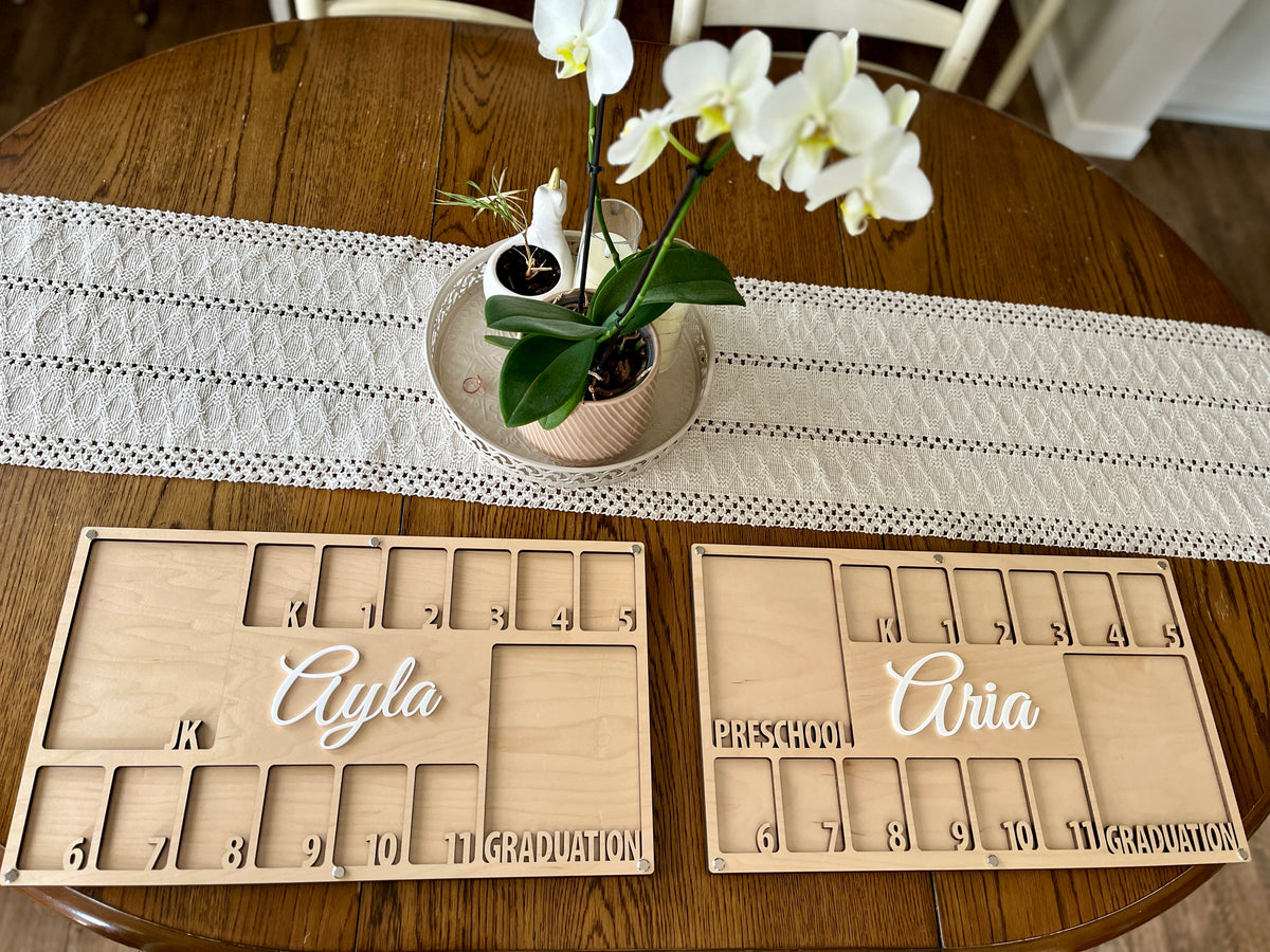 Two wooden milestone boards with engraved numbers and white script names displayed on a wooden table with a lace runner and white orchids centerpiece