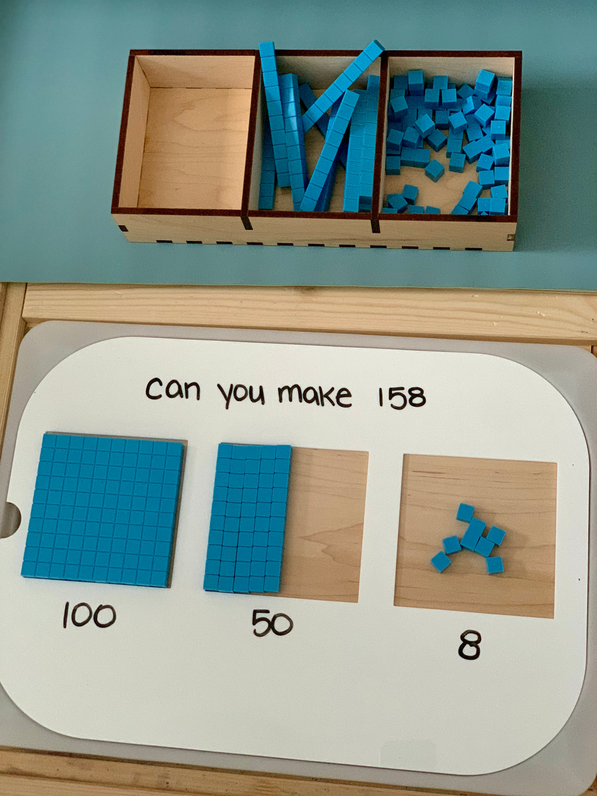 Wooden math base ten blocks arranged on a whiteboard solving 158 with hundreds, tens, and ones blocks