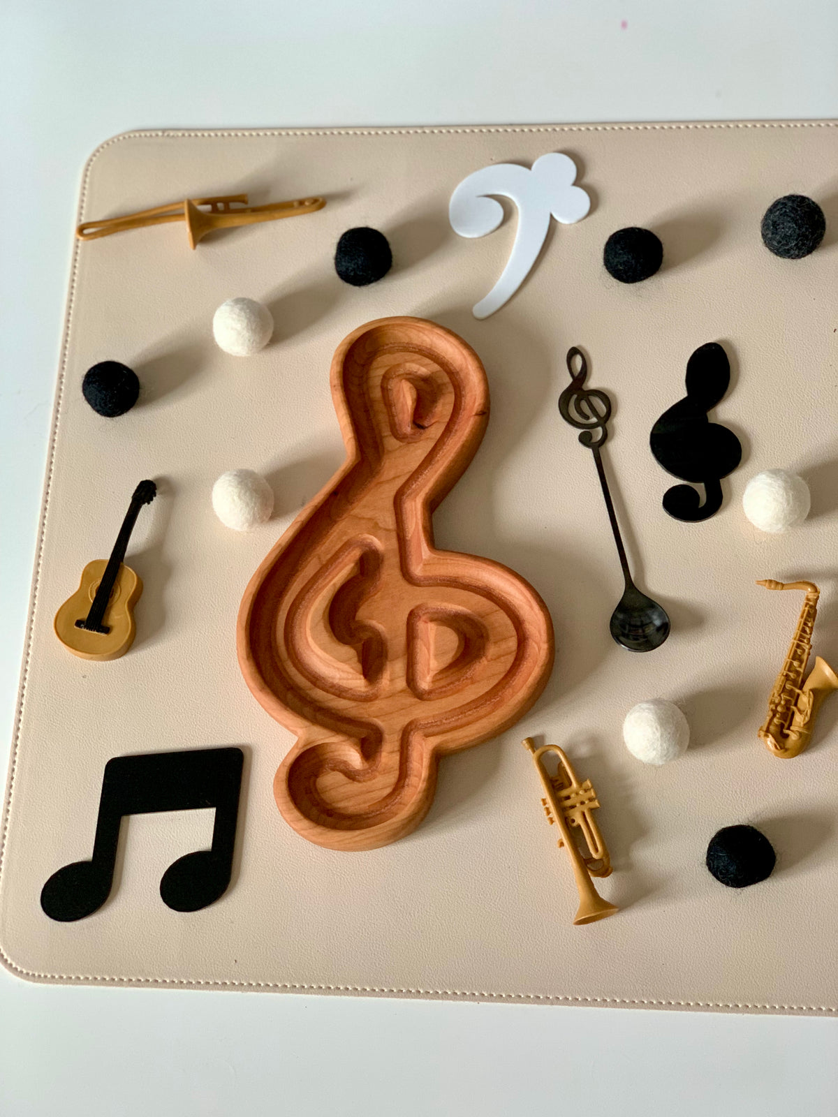 Wooden treble clef tray surrounded by black and white felt balls, miniature musical instruments, and black music note cutouts on beige surface