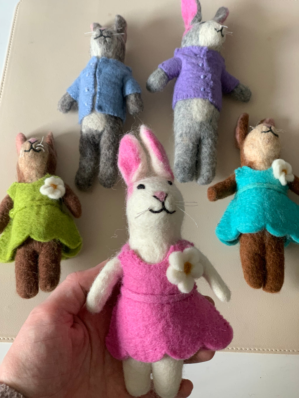 Five handcrafted felt animal dolls dressed in colorful outfits on a beige surface with one held in hand