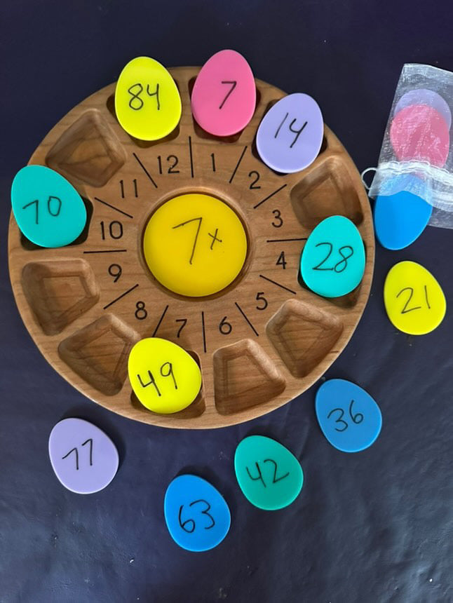 Round wooden multiplication board with colorful numbered egg-shaped tiles arranged and some scattered on a dark surface