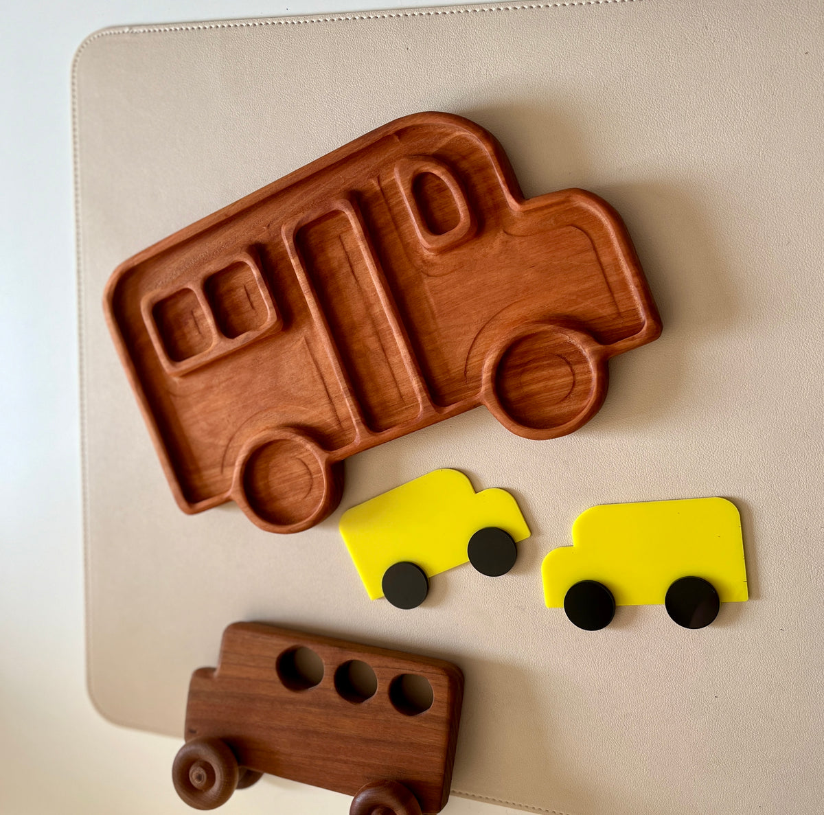 Wooden tray shaped like a camper van with two small yellow toy vans and a dark wooden bus toy on a light surface