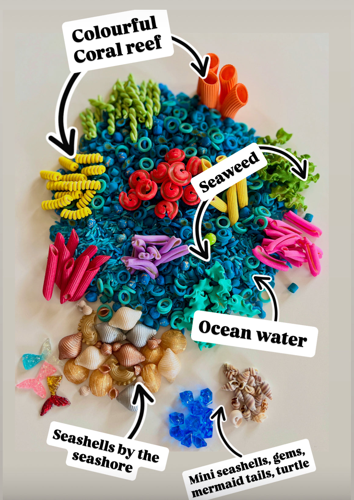 Assorted colorful craft pasta shapes, seashells, and blue beads arranged to mimic ocean coral reef and sea elements