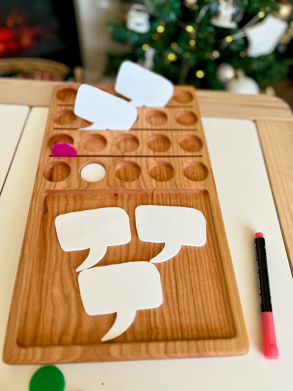 Wooden game board with blank white speech bubble cutouts, colored game pieces, and a pink marker on white surface
