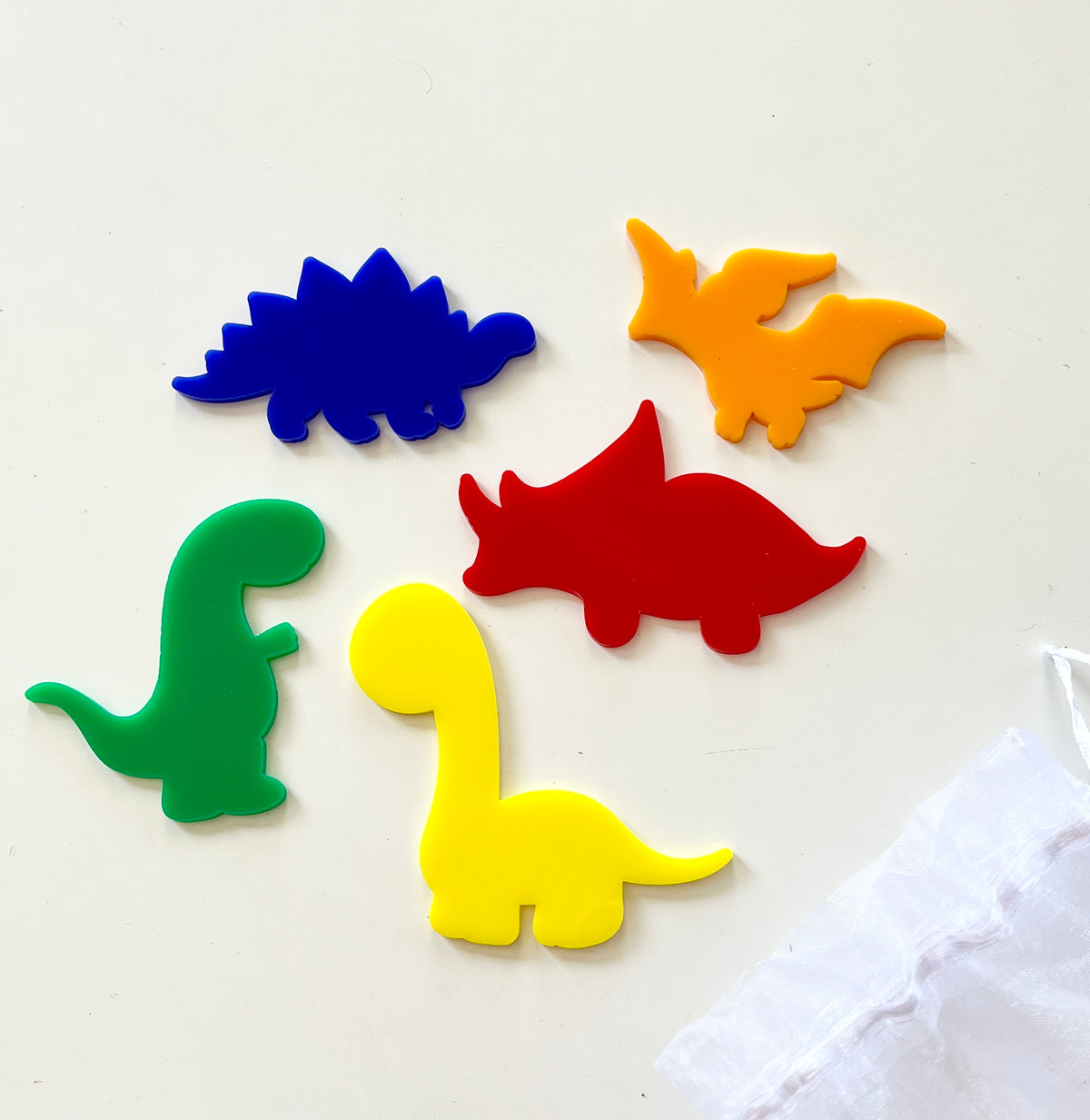 Five colorful acrylic dinosaur shapes including blue stegosaurus, orange pterodactyl, red triceratops, green T-rex, and yellow brachiosaurus on white surface