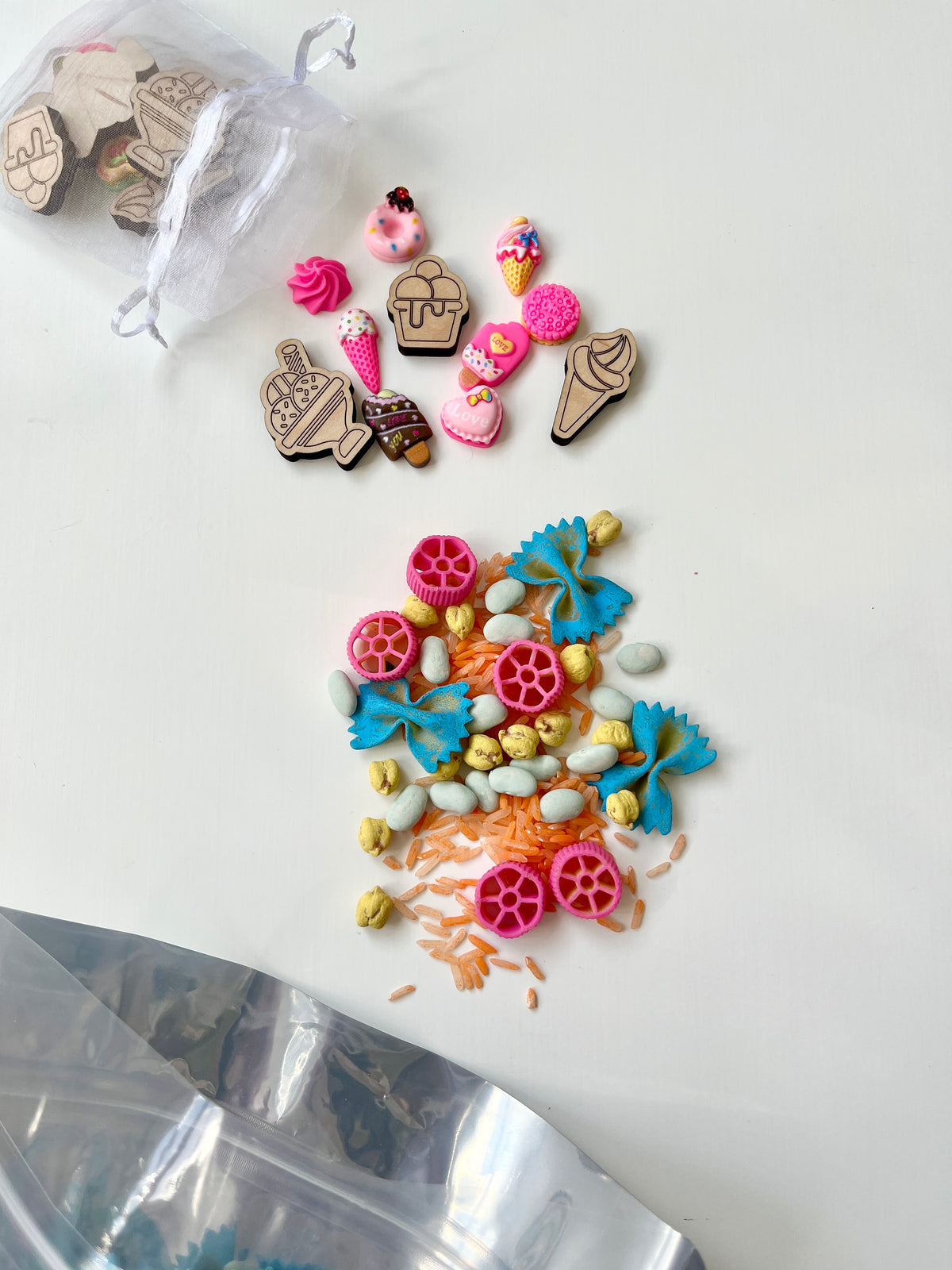 Small wooden dessert and ice cream shapes scattered near blue, pink, and white pasta pieces on white surface with drawstring bag above.