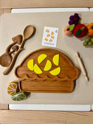 Wooden play food set with a muffin-shaped tray holding six yellow lemon wedges and surrounded by toy cooking utensils and felt fruit.