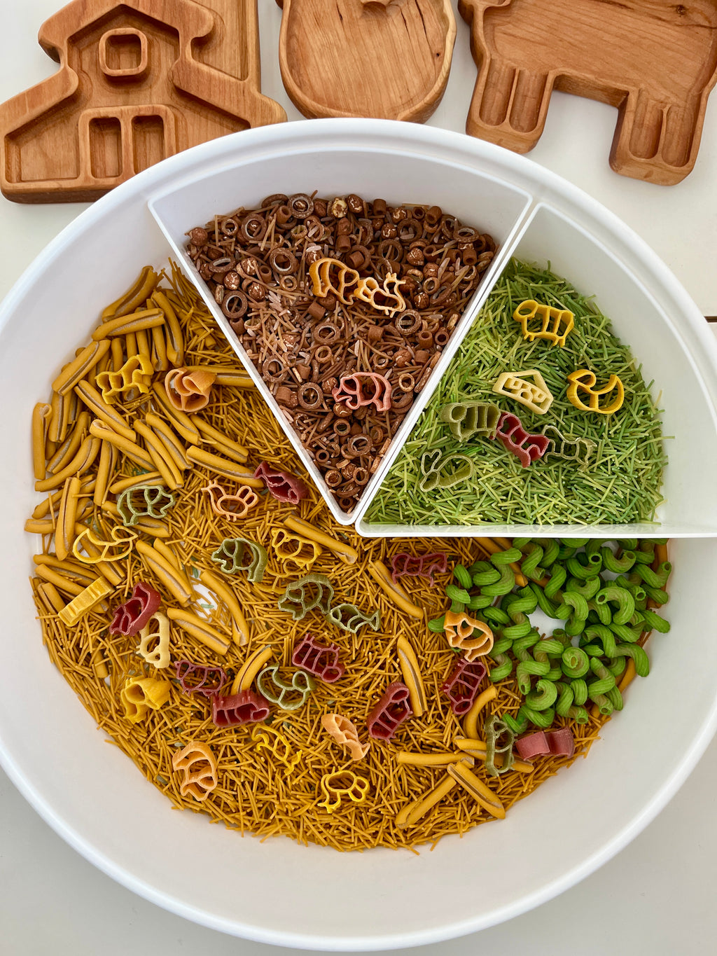 White segmented tray with green, brown, yellow pasta and small animal-shaped wooden trays on white surface