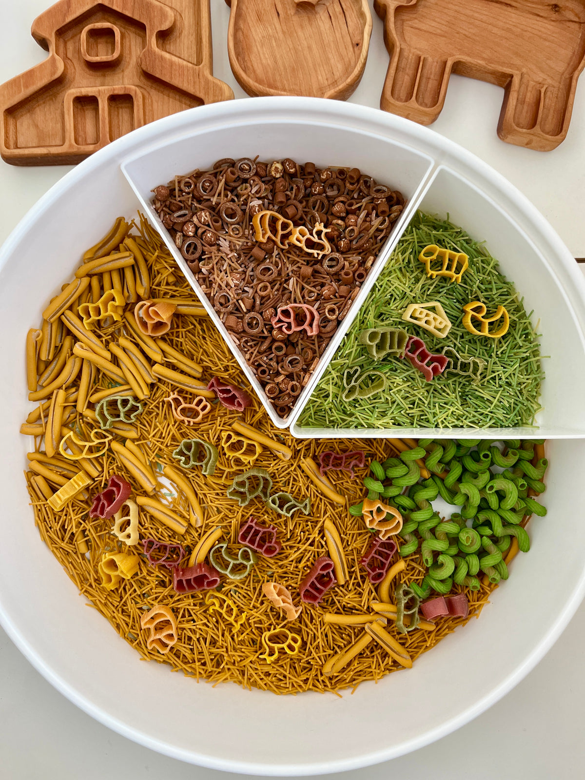 White segmented tray with green, brown, yellow pasta and small animal-shaped wooden trays on white surface