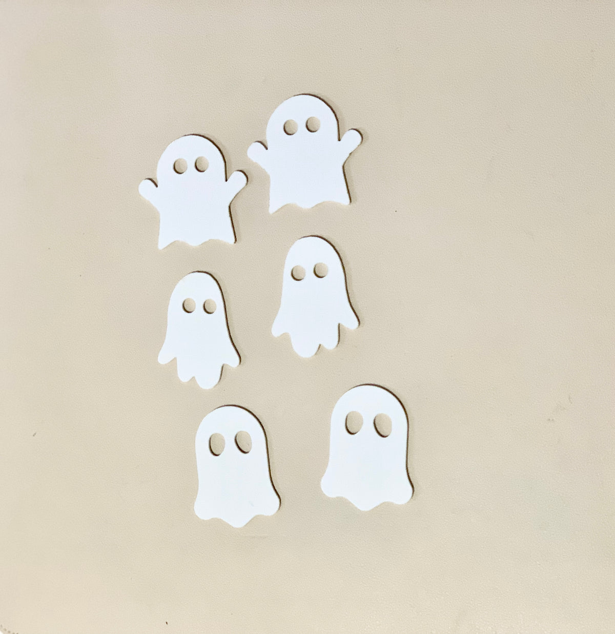 Six white wooden ghost shapes with cut-out eyes arranged on a beige background