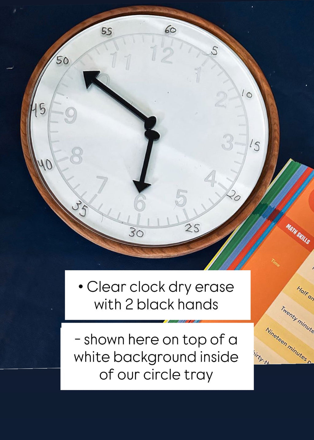 Round wooden-framed dry erase clock with black hands on dark surface next to colorful learning cards