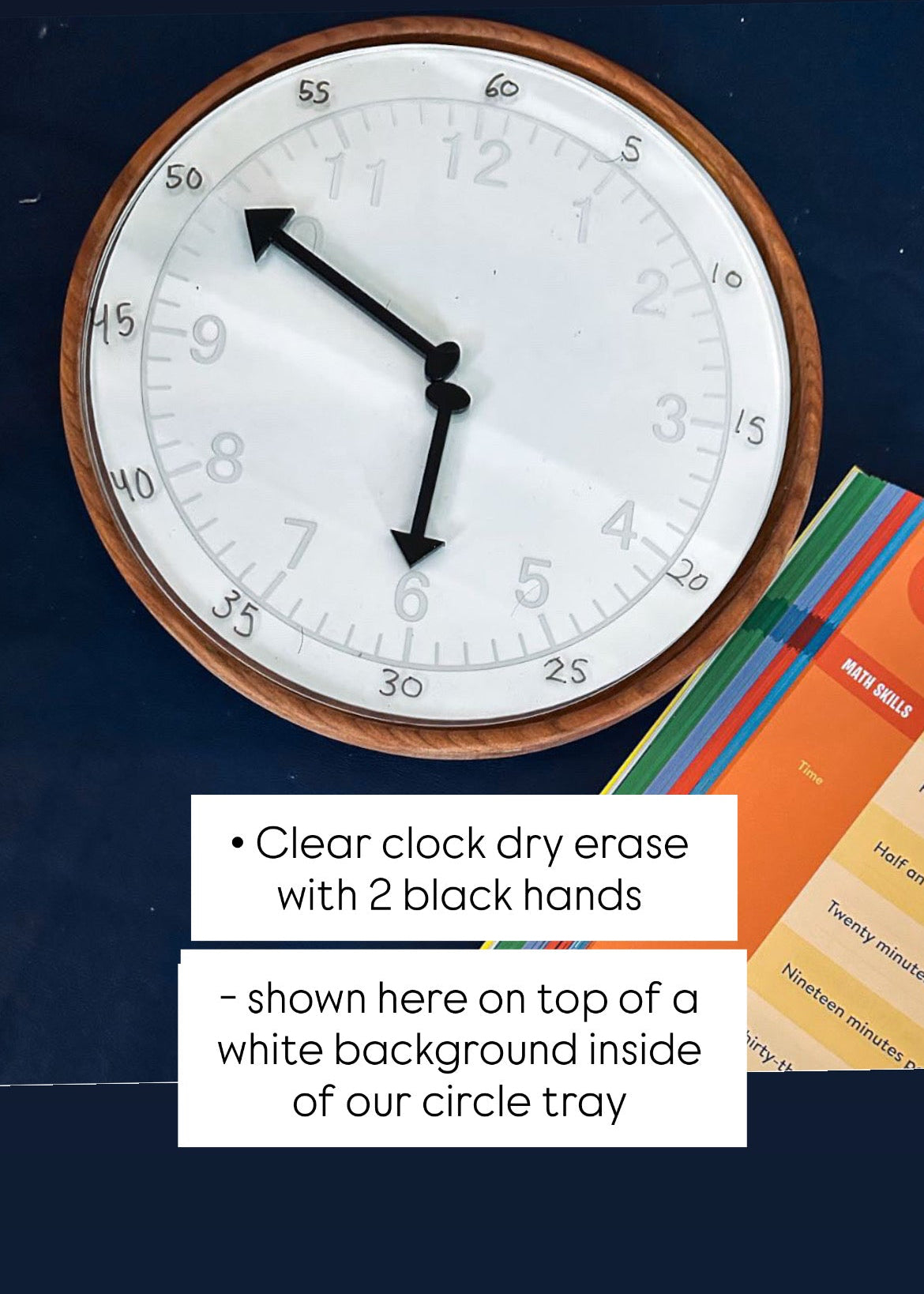 Round wooden-framed dry erase clock with black hands on dark surface next to colorful learning cards