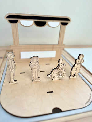 Wooden laser-cut figures including a person in a wheelchair, two standing people, and a street vendor awning on a light wood base