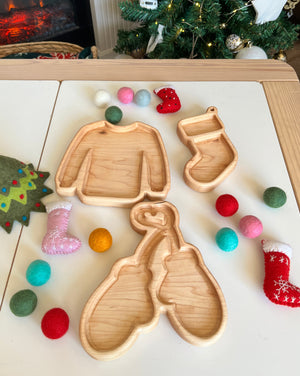 Wooden trays in sweater, mitten, and stocking shapes surrounded by colorful felt balls and small holiday stockings on table
