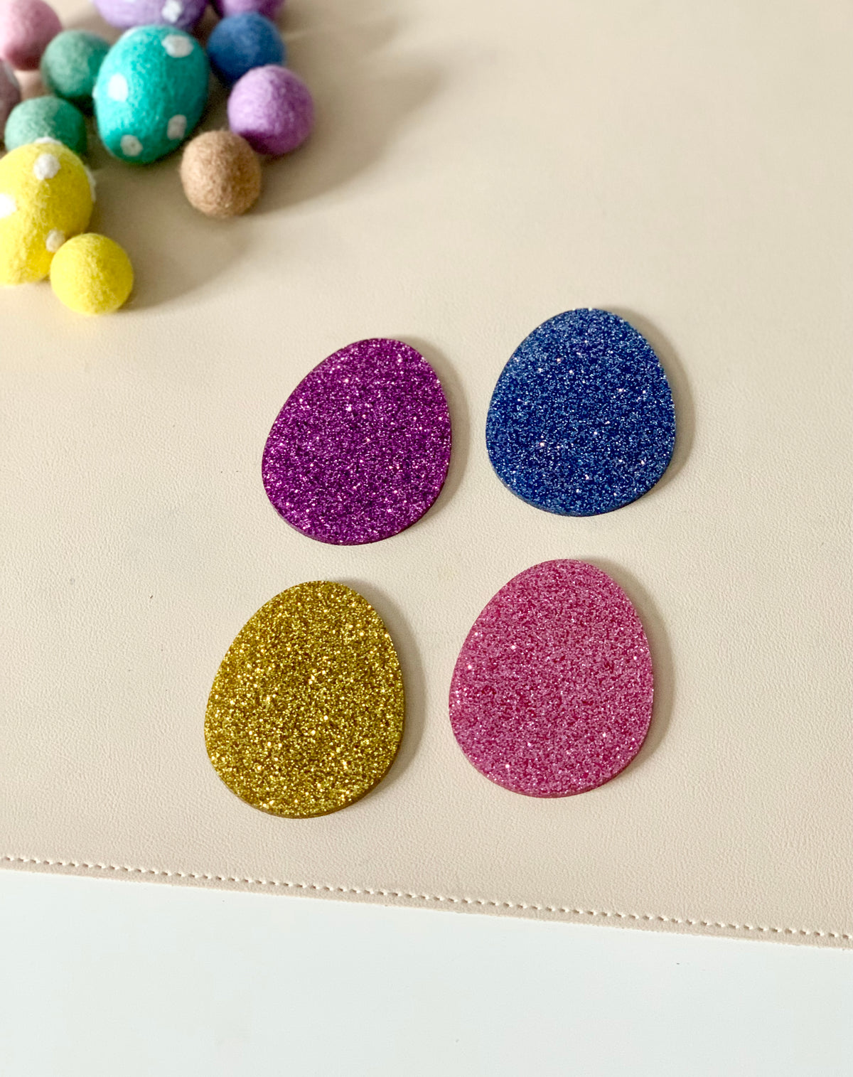 Four glittery egg-shaped cutouts in purple, blue, gold, and pink on a light beige surface with felt balls nearby