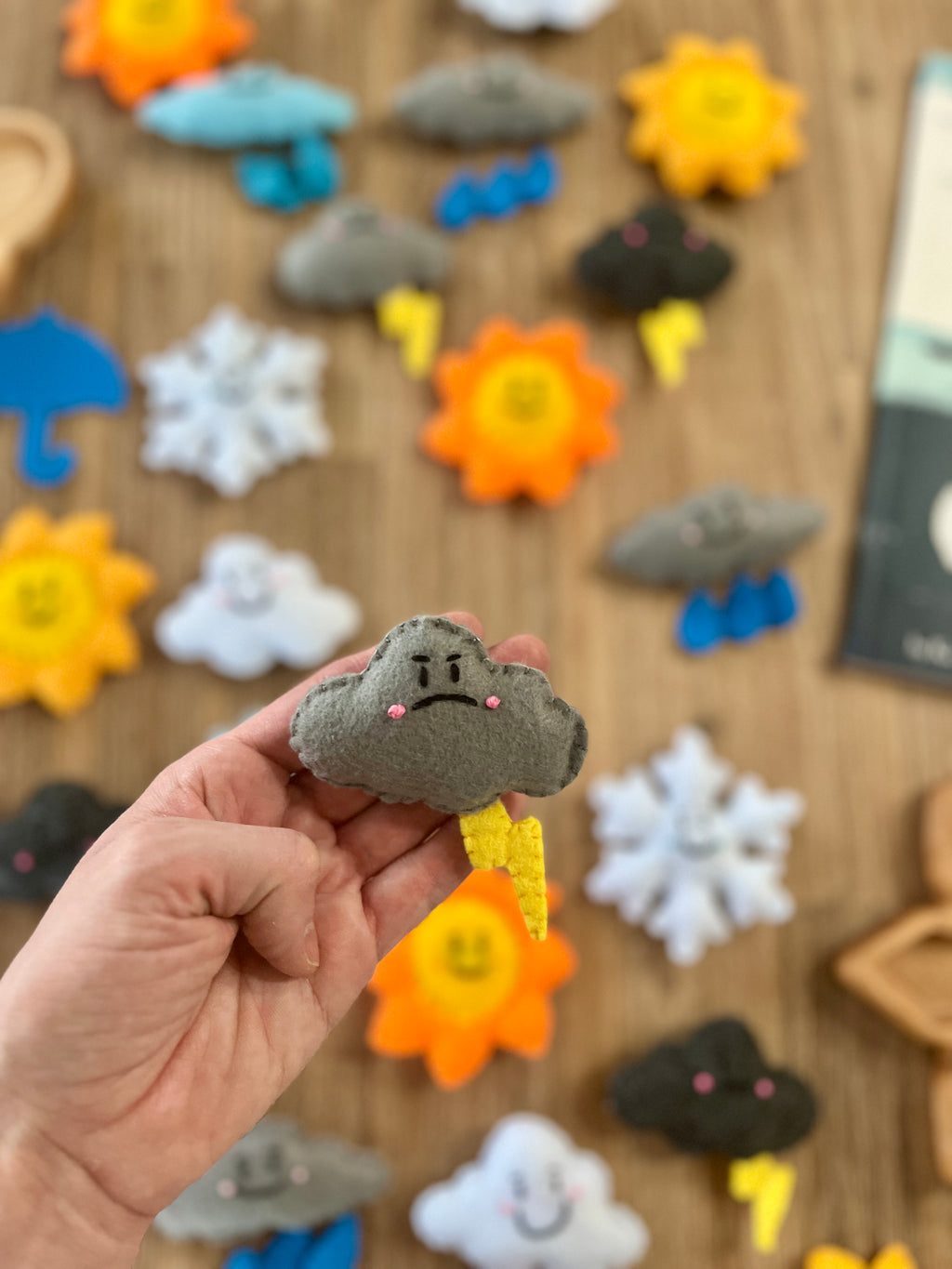 Hand holding a gray felt cloud with an embroidered angry face and yellow lightning bolt, with other colorful Felt Weather Shapes, Made in Canada, handcrafted, homeschool, education resources scattered on a wooden surface.