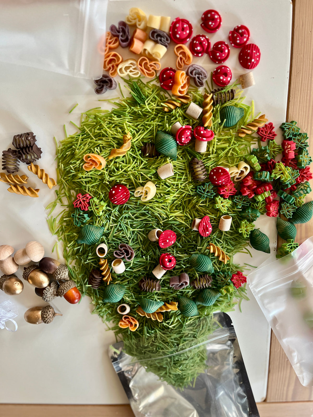 Assortment of colorful pasta shapes and wooden acorns spread on a white surface with some plastic bags nearby