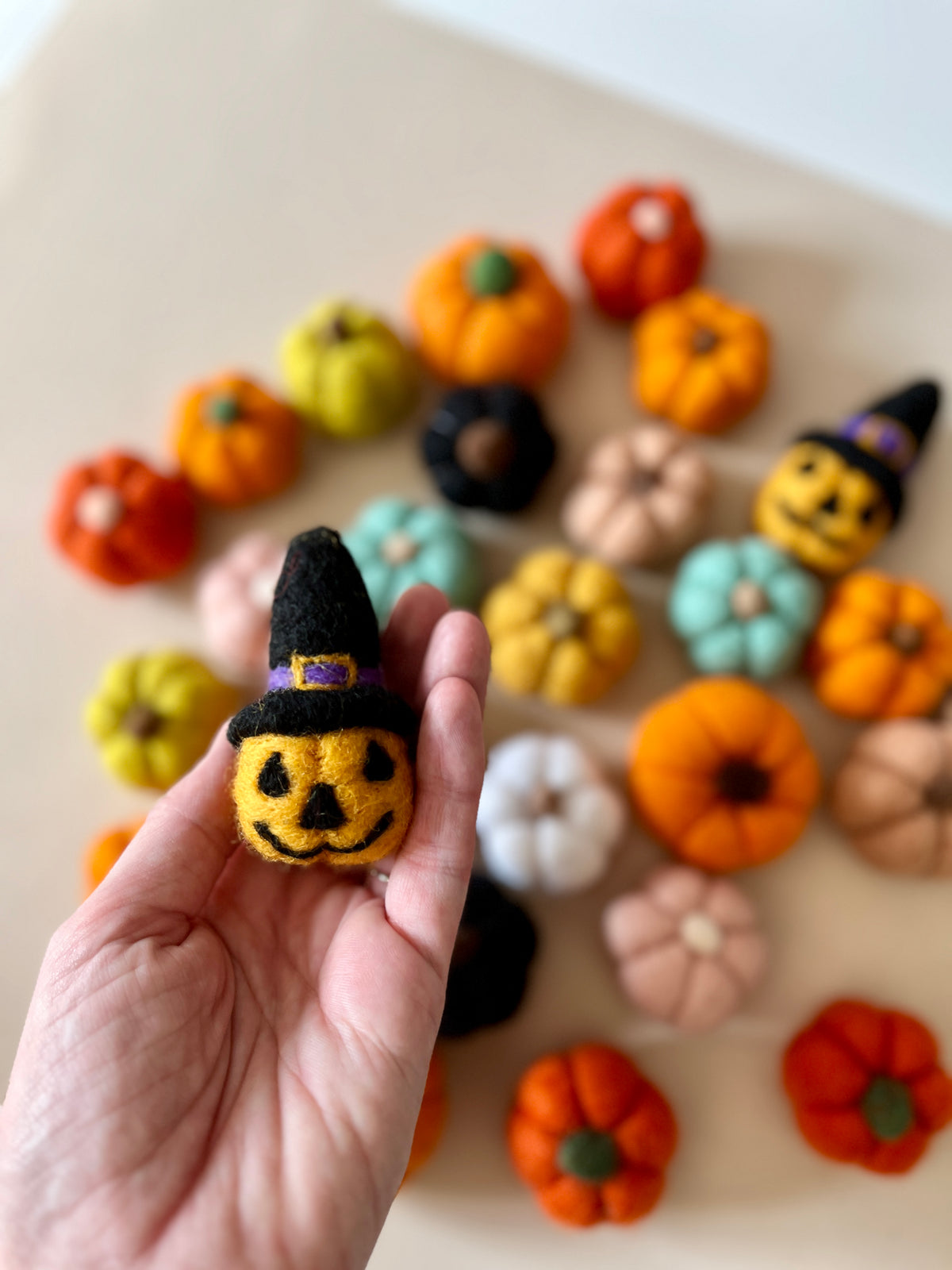 Hand holding a small felt jack-o-lantern wearing a black witch hat with various colorful felt pumpkins on a beige background