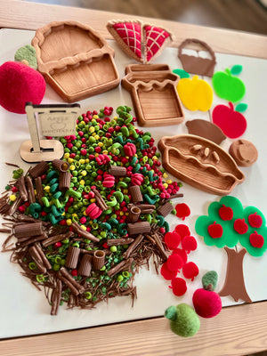 Miniature wooden trays and felt apple craft pieces arranged with colorful pasta and twigs on a light surface
