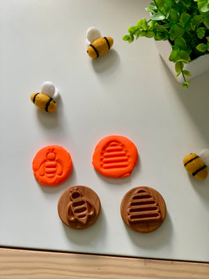 Wooden stamps with bee and hive designs pressed into orange clay, surrounded by three felted bee toys and a green plant on white surface