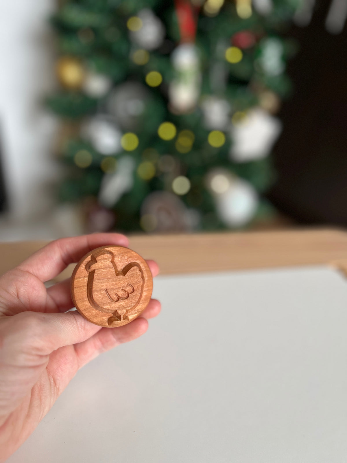 Hand holding round wooden stamp with carved hen design against blurred holiday-themed background
