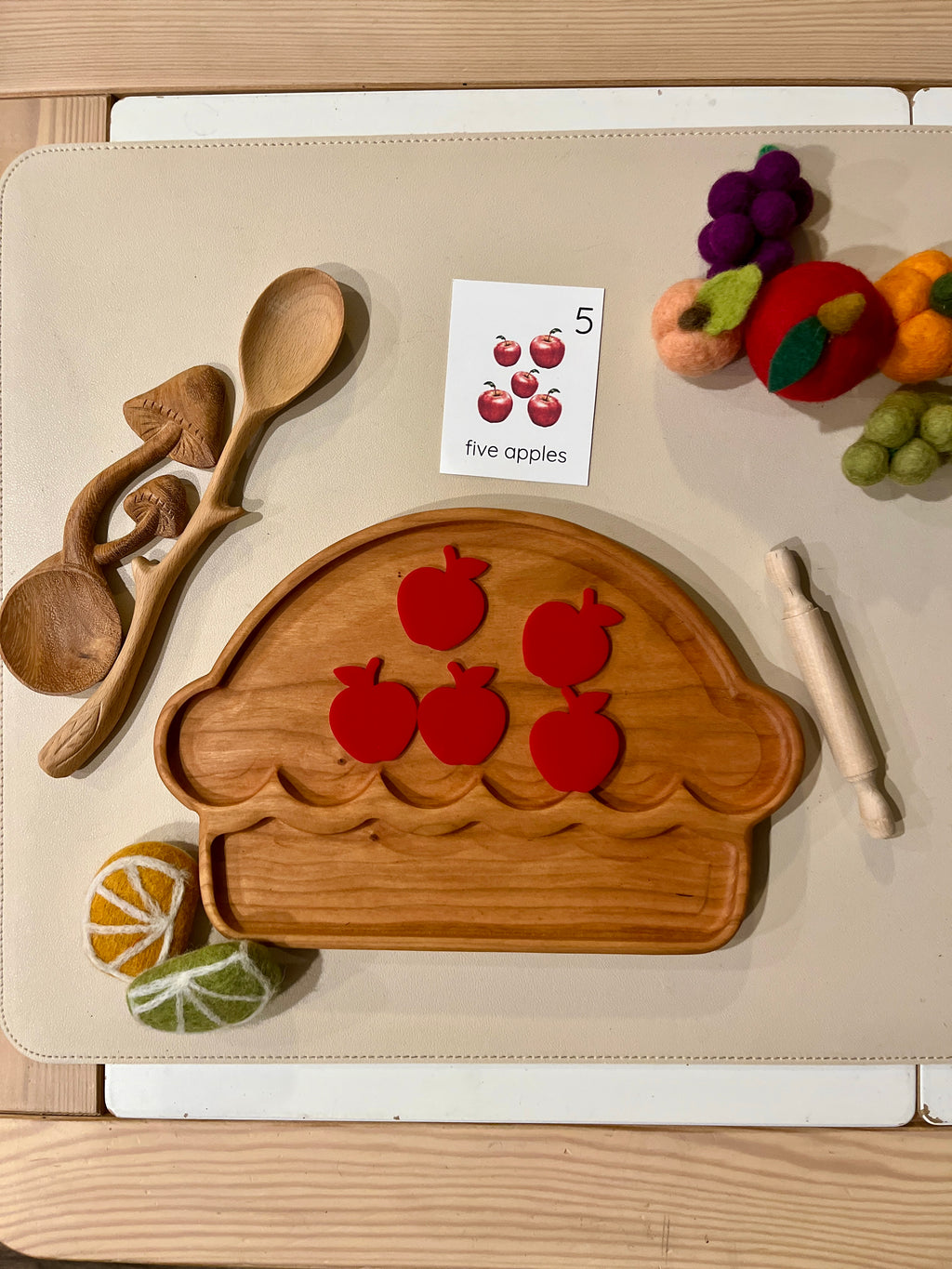 Wooden cupcake-shaped tray with five red apple cutouts, wooden spoons, rolling pin, and felt fruit on a beige surface