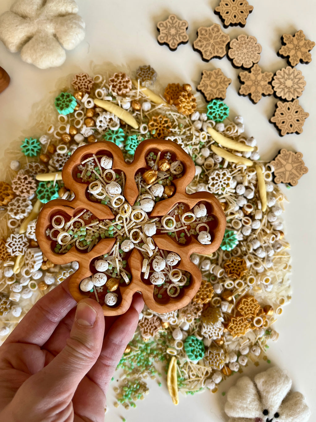 Hand holding wooden tray shaped like a flower filled with small decorative winter-themed pieces on a white surface