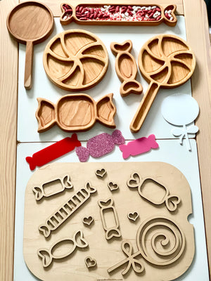 Wooden trays shaped like candies and lollipops with red and white beans, colorful paper cutouts, and small hearts on light background