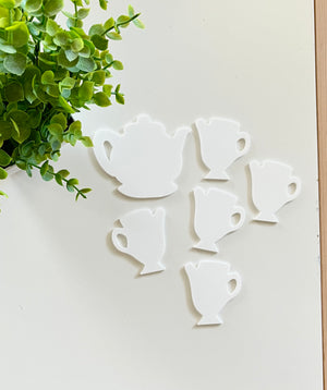White tea pot and tea cup-shaped cutouts arranged on white surface near small green leafy plant