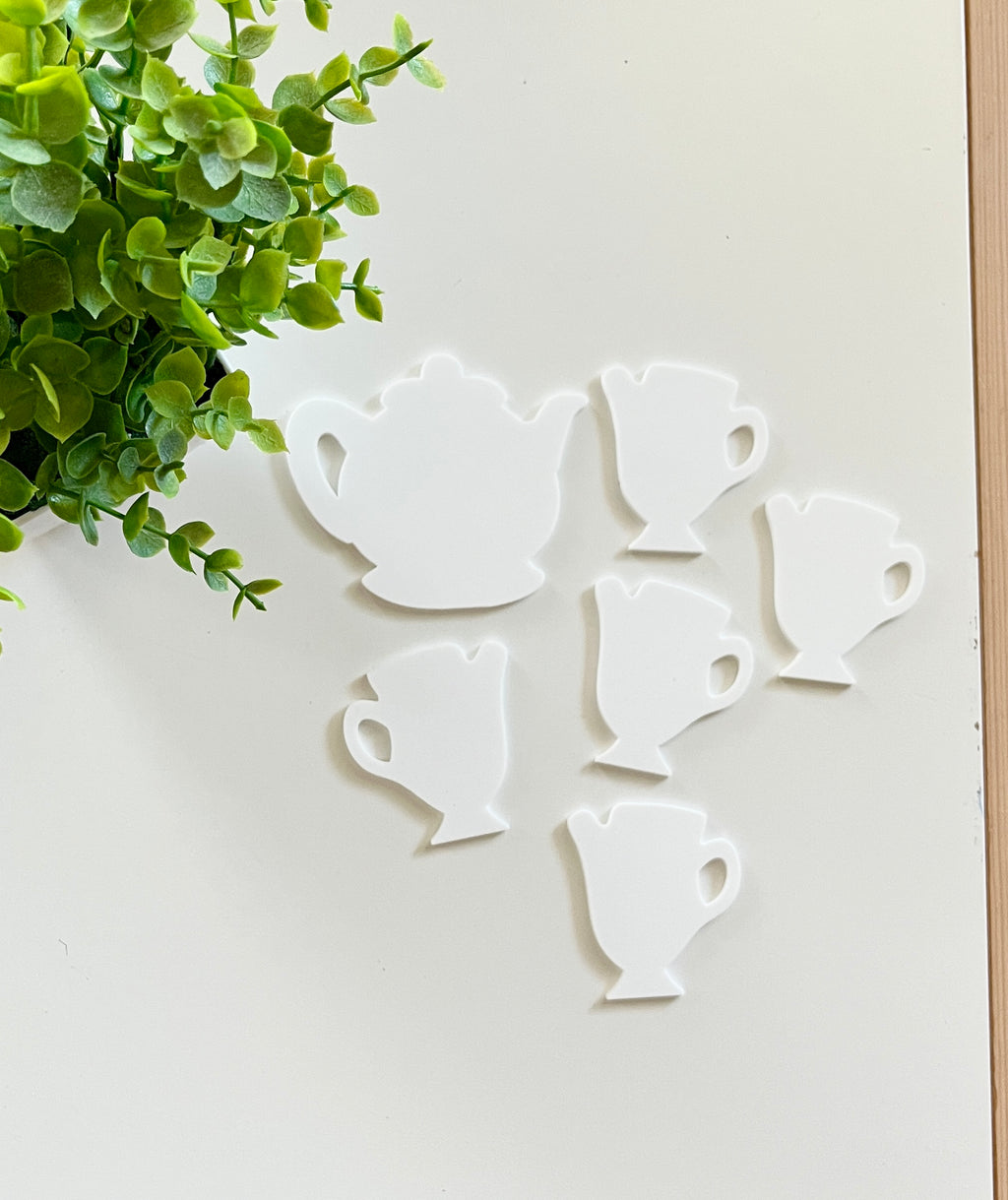 White tea pot and tea cup-shaped cutouts arranged on white surface near small green leafy plant