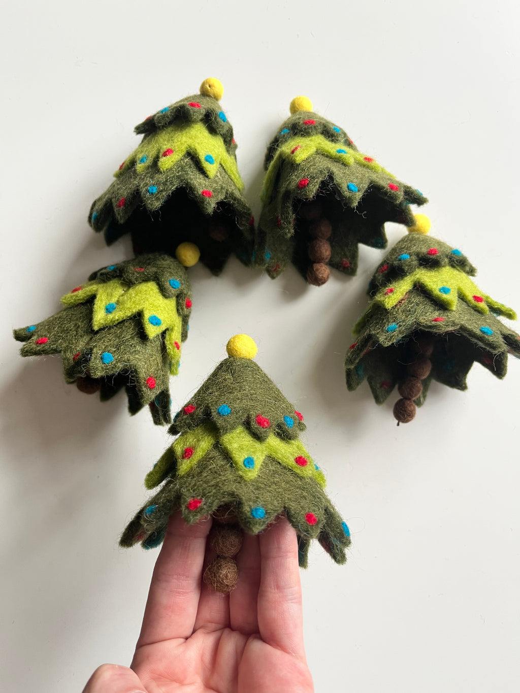 Small handmade felt Christmas trees in shades of green with colorful round ornaments and yellow felt tops held on fingers and on white surface