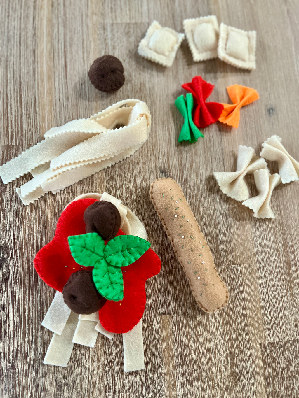 Various felt pasta shapes including beige noodles, red and orange farfalle, ravioli, meatballs, and a baguette, arranged on a wooden surface, Felt Pasta Set, Made in Canada, handcrafted, homeschool, education resources.