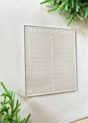Clear acrylic alphabet game board with engraved letters arranged in two columns on light surface with green foliage accents
