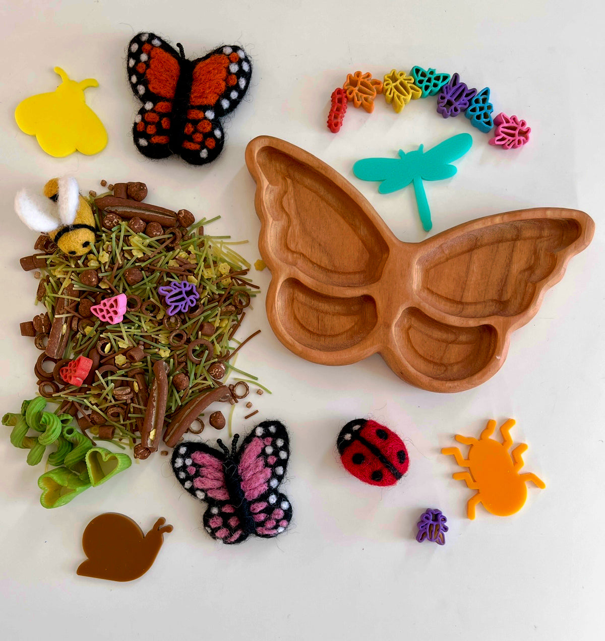 Wooden butterfly-shaped tray surrounded by felt insects, plastic bug shapes, dried natural materials, and colorful craft pieces on white background