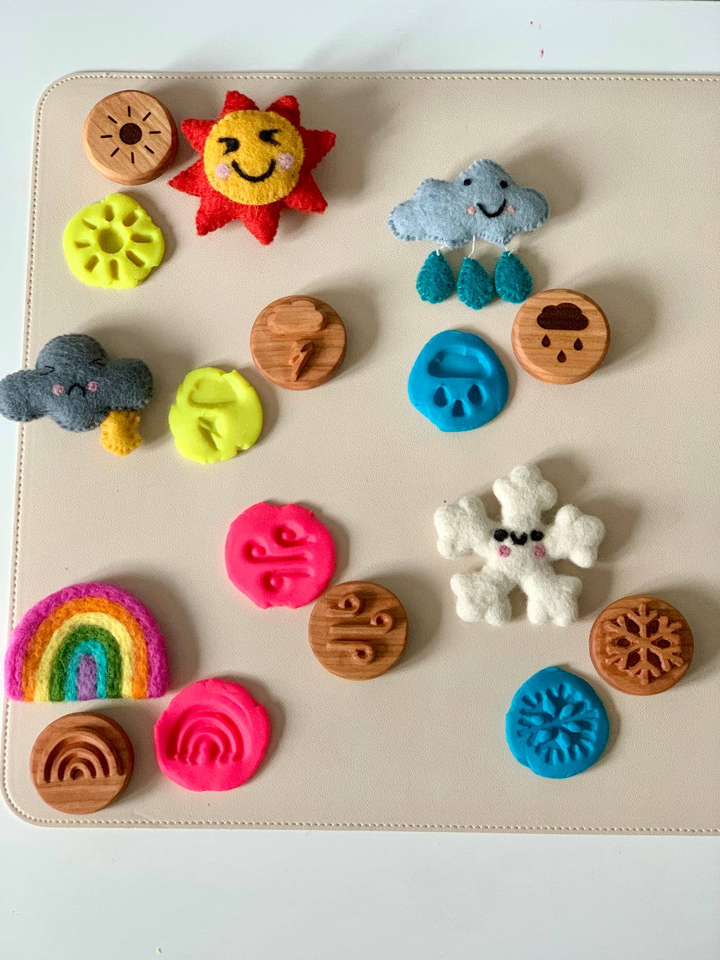 Assorted wooden and felt weather symbols including sun, clouds, rainbow, snowflake, and colorful textured clay shapes on a light surface