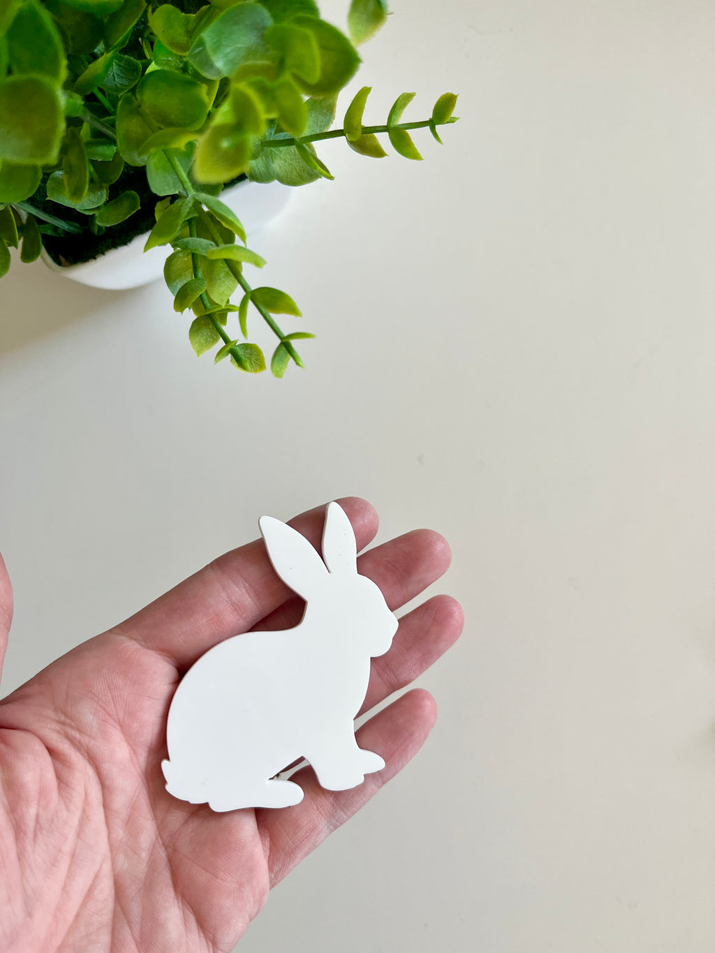 Hand holding a smooth white rabbit-shaped cutout with a green leafy plant in a white pot visible above