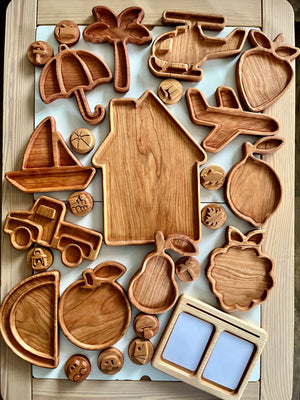 Wooden tray shaped like a house surrounded by carved trays and round tokens including airplane, palm tree, fruit, and vehicle designs on white surface