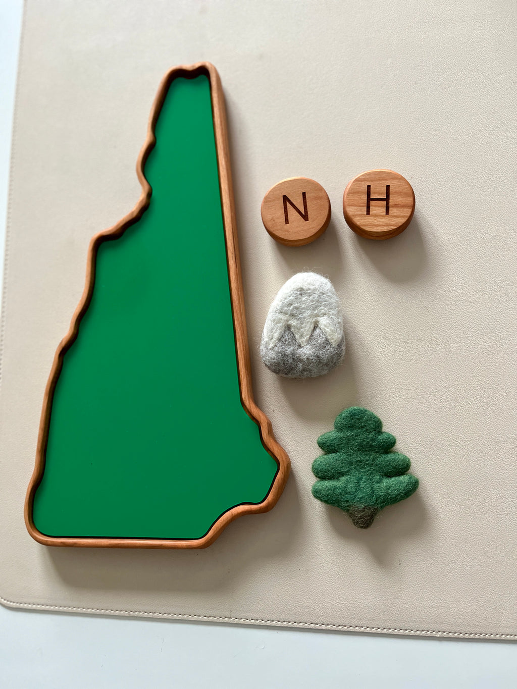 Wooden tray shaped like New Hampshire with felt mountain, pine tree, and two wooden letter blocks on beige surface