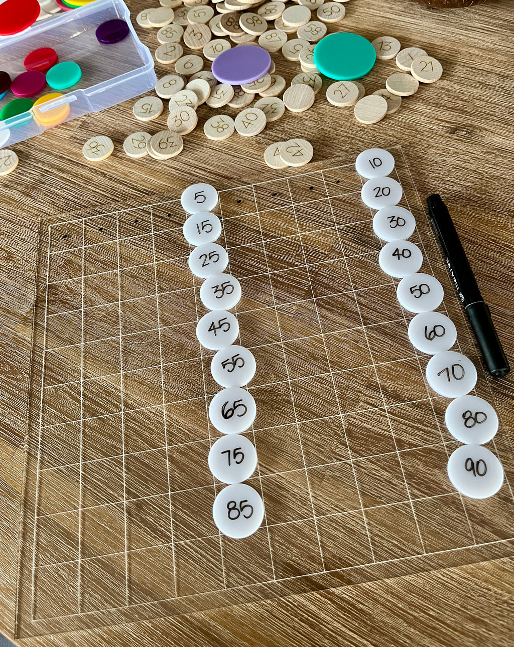 Dry Erasable Blank Open Ended 100 Frame Hundred Board on wood table with translucent grid and numbered white discs arranged in columns, black dry erase marker, handcrafted homeschool education resources.