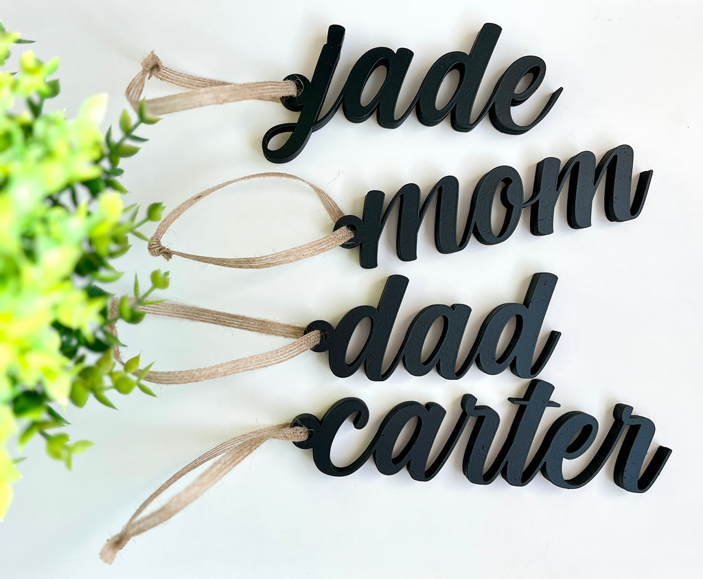 Four black cursive name tags reading jade, mom, dad, and carter with tan hanging ribbons on white surface
