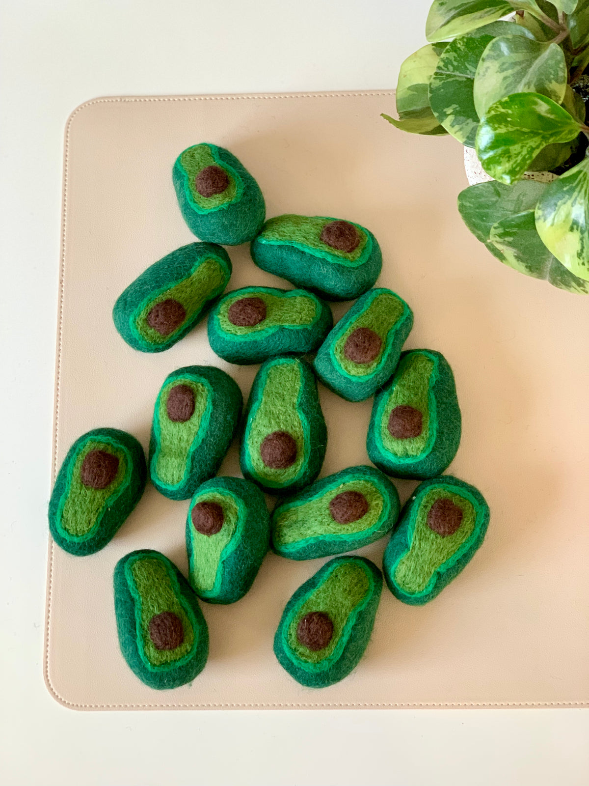 Group of small felted wool avocados with brown pits arranged on a beige surface near green houseplant leaves