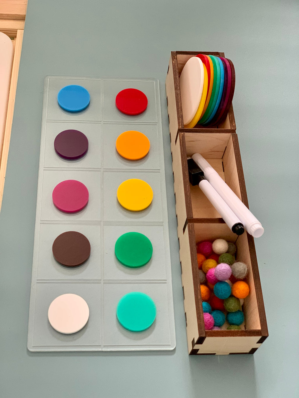 Clear plastic grid displaying colorful round magnets beside wooden box holding more magnets, felt balls, and two white pens