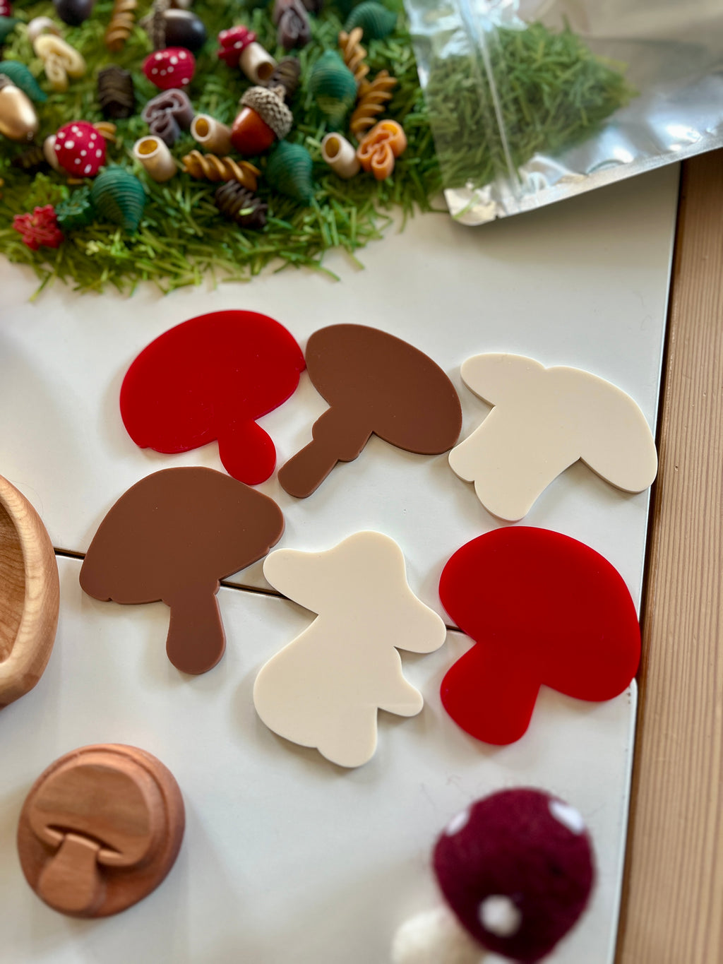 Assorted red, brown, and white mushroom-shaped cutouts on white tiles with craft supplies and greenery nearby