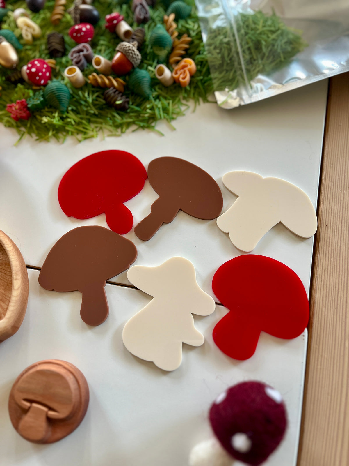 Assorted red, brown, and white mushroom-shaped cutouts on white tiles with craft supplies and greenery nearby