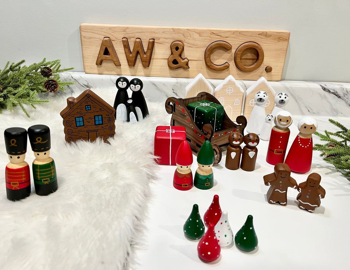 Wooden holiday figurines including soldiers, gnomes, gingerbread people, penguins, polar bears, sleigh, gift box, and houses on marble surface