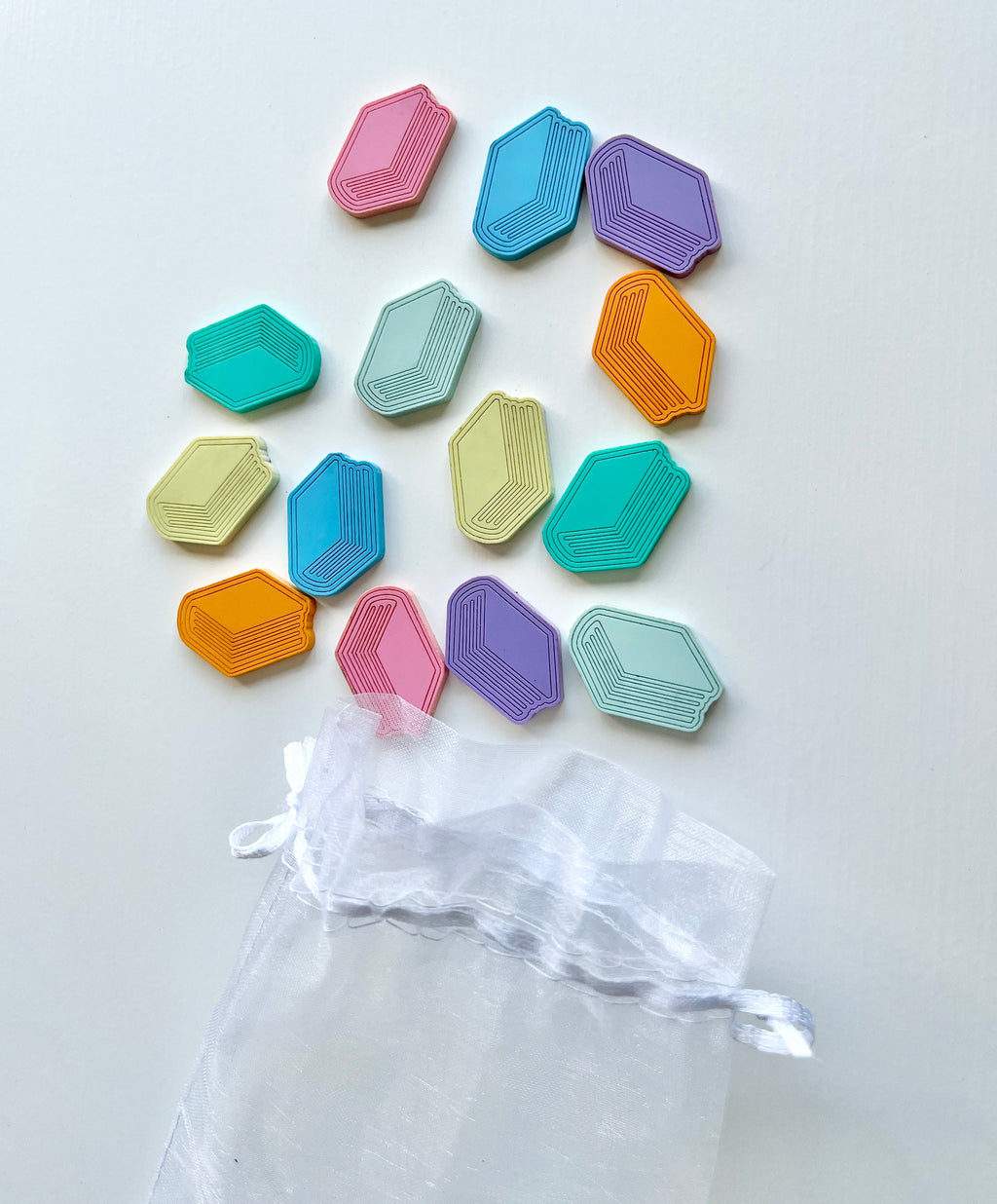 Multicolored geometric game tokens scattered above a translucent white drawstring pouch on a white background