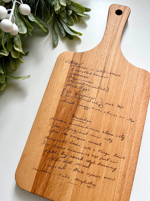 Natural wooden personalized cutting board with laser engraved handwritten recipe and leafy green decoration on white surface