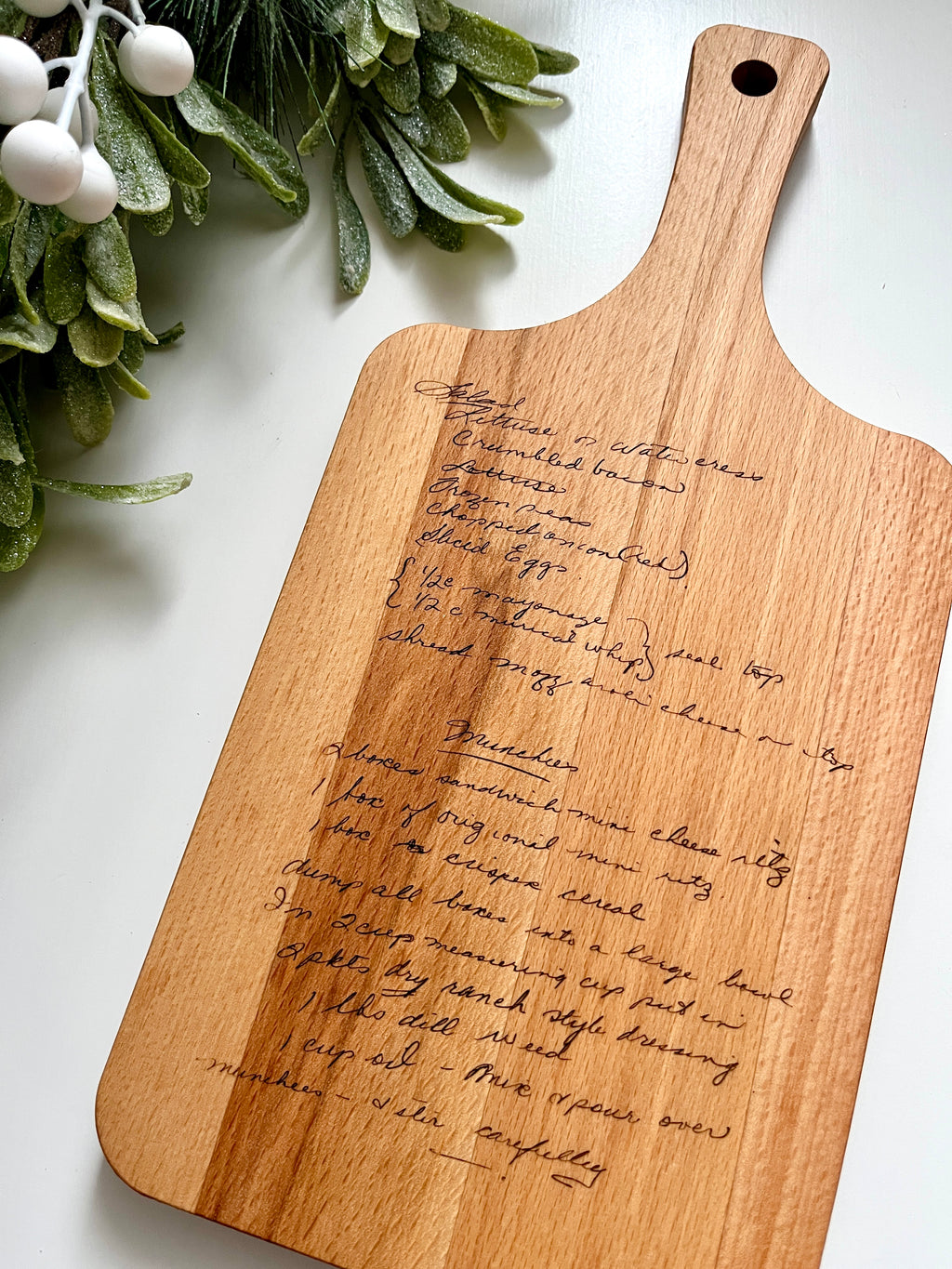Natural wooden personalized cutting board with laser engraved handwritten recipe and leafy green decoration on white surface